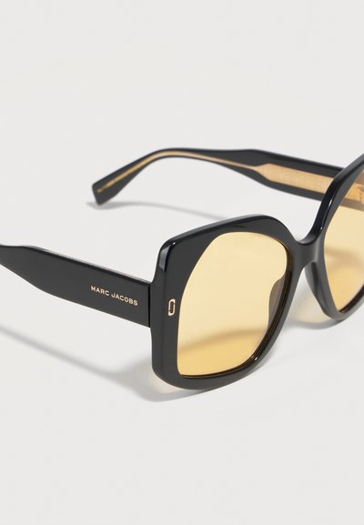 Μαύρα γυαλιά ηλίου Marc Jacobs με χοντρά τετράγωνα πλαίσια και κίτρινα φακούς σε λευκό φόντο.