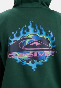 Sudadera verde oscuro con un logo colorido de Quiksilver que muestra olas y llamas estilizadas en azul, amarillo y rosa en la parte posterior.