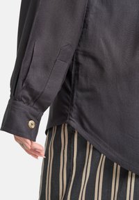 Camisa de color gris oscuro, con cuello y mangas largas, que presenta puños abotonados, tejido texturizado y dobladillo lateral inclinado, combinada con pantalones de rayas verticales.