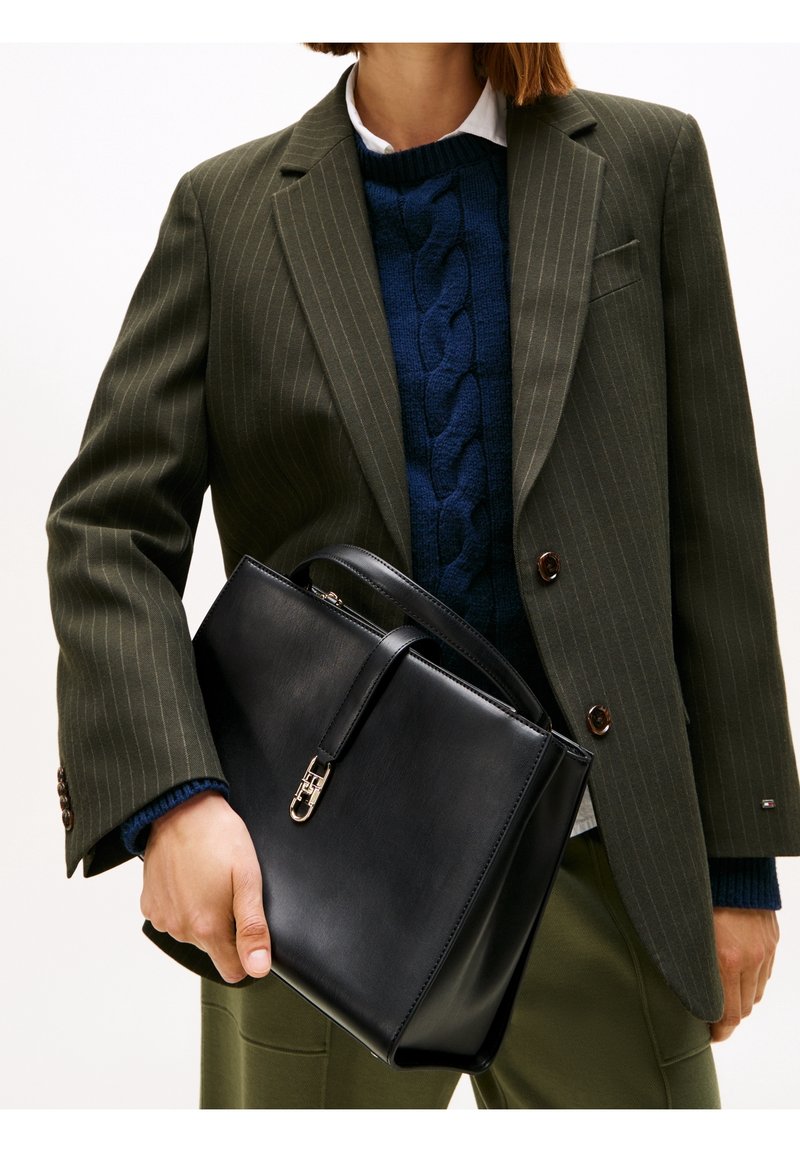 Personne portant un blazer à fines rayures vert foncé et un pull bleu marine, tenant un sac à main en cuir noir avec un fermoir doré devant un fond blanc.