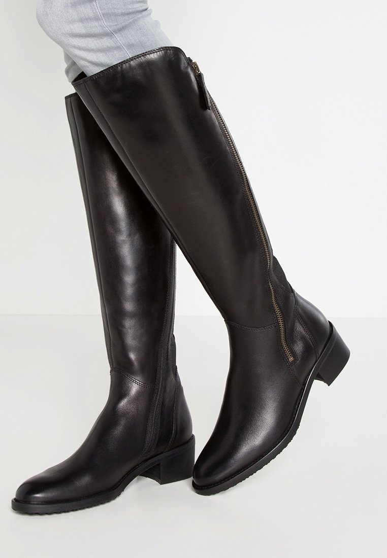Bottes en cuir noires montantes jusqu'aux genoux avec fermeture éclair latérale portées avec un jean bleu clair sur fond blanc.