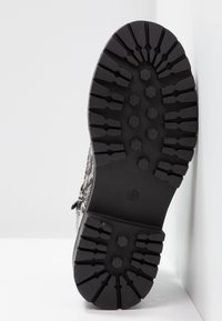 Semelle de botte noire avec des motifs de bande de roulement hexagonaux profonds et linéaires, inclinée contre un mur et un sol blancs.