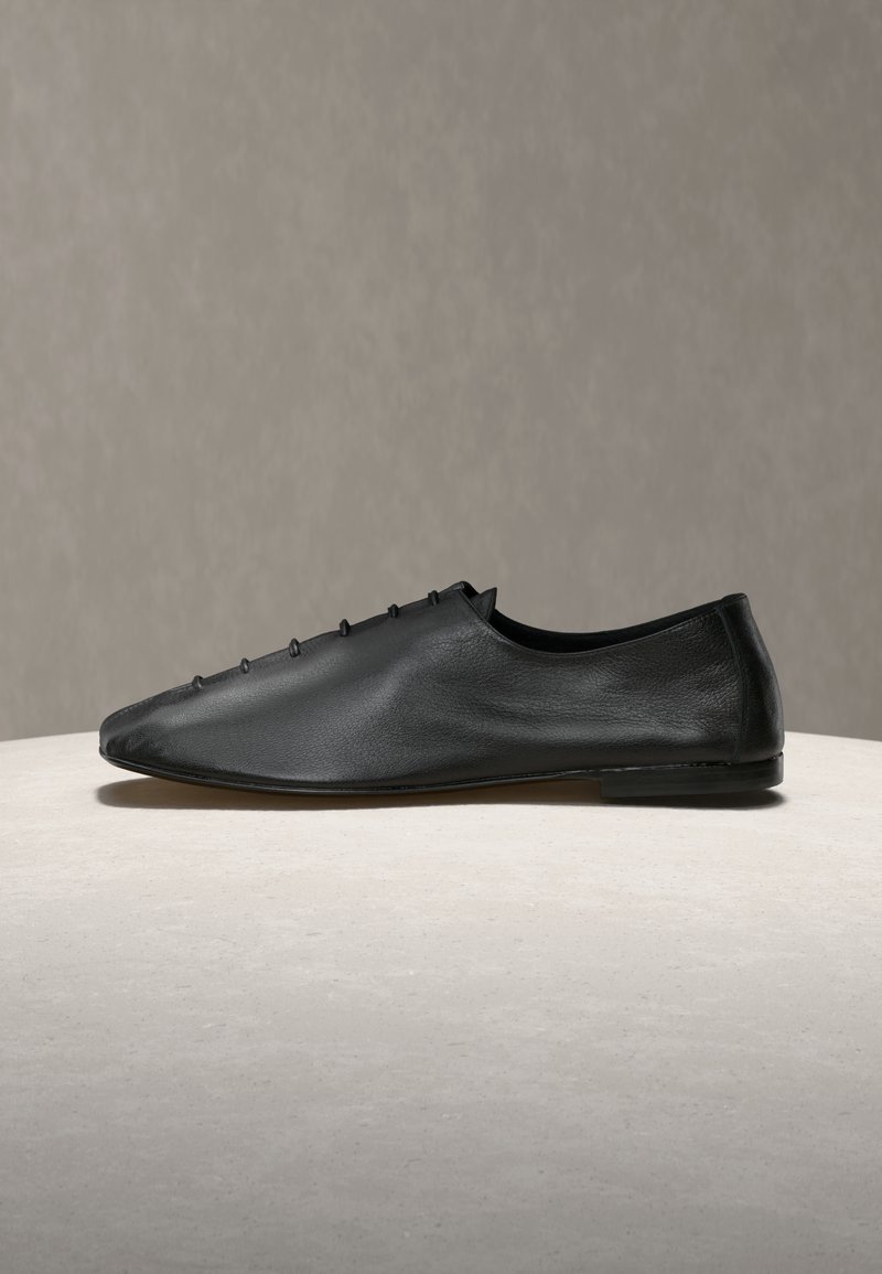Črni usnjeni čevlji brez vezalk z zašiljenim prstom, z majhnimi okrasnimi žeblji na zgornjem delu in gladkim, mat zaključkom.