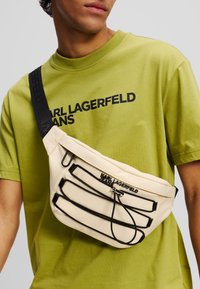 Beige Stoff Umhängetasche mit schwarzen Akzenten, ausgestattet mit drei Reißverschlussfächern und einem Logo. Getragen über einem grünen Shirt.