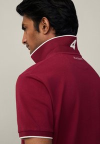 Burgundská polo košeľa s prevráteným golierom, bielymi akcentmi a vyšitým číslom '4' na zadnej strane. Bavlnený materiál s textúrovaným povrchom.