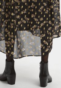 Abito nero con stampa floreale in tessuto trasparente, caratterizzato da motivi floreali beige e oro. Abbinato a stivali neri alla caviglia e collant opachi.