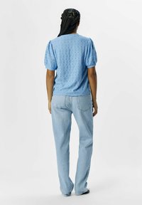 Ljusblå blus med broderade hål och puffärmar, matchad med ljusa denimjeans. Blusen har en texturerad design och rundad halsringning.