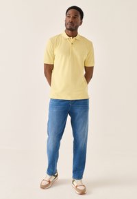 Gul kortærmet polo-shirt med krave, med en lille logo på brystet, kombineret med blå jeans med lige ben og flerfarvede sneakers.