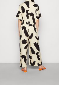 Monos de estampado negro y crema con mangas cortas, piernas anchas y un ajuste relajado. Presenta una cintura ceñida y sandalias tipo slides en color naranja.