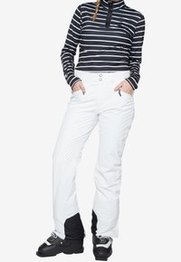 Donna che indossa un top a righe bianche e nere con maniche lunghe, pantaloni da sci bianchi con tasche con zip e scarponi da sci neri, in piedi con le mani nelle tasche.