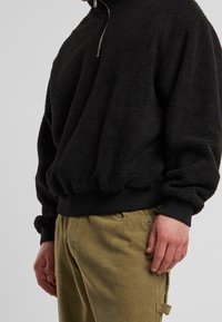 Sort fleece-pullover med høj krave og en sølvlynlås. Har ribbede manchetter og kant, struktureret stof, båret over lysfarvede fløjlshorts.
