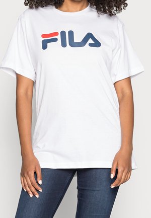 Osoba ubrana w białą koszulkę FILA z granatowo-czerwonym logo, w połączeniu z ciemnoniebieskimi dżinsami, na jednolitym jasnym tle.