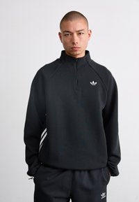 Sweat à zip noir avec col haut et logo Adidas blanc. Comprend des manches raglan et trois bandes blanches sur les côtés.