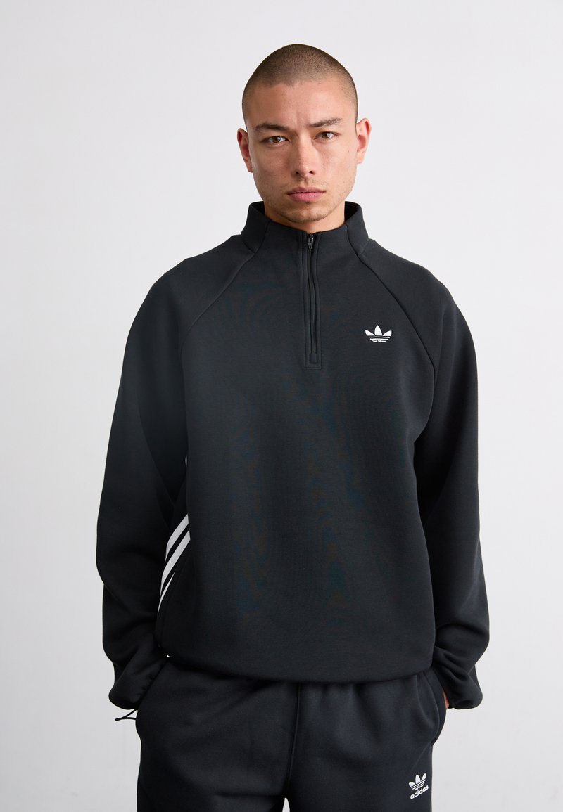 Sweat à zip noir avec col haut et logo Adidas blanc. Comprend des manches raglan et trois bandes blanches sur les côtés.