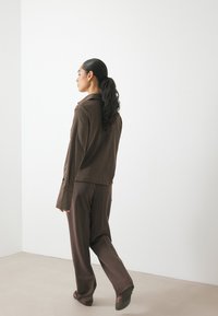 Brown textured jacket with a collar, long sleeves, and buttoned cuffs, paired with dark brown straight-leg trousers and simple footwear.