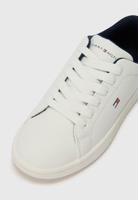Beli športni copat iz gladkega usnja z okroglo konico, vezalkami in mornarsko modrim notranjim delom. Ima majhen grb z rdečimi in modrimi akcenti.