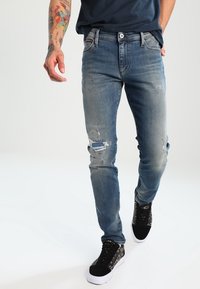 Jeans de mezclilla azul con corte Slim, detalles desgastados y un diseño clásico de cinco bolsillos. Combinados con zapatillas negras con un acento estampado.