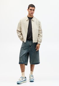 Beige bomberjack, zwarte t-shirt, oversized denim shorts en sneakers met blauwe accenten. Effen achtergrond, geen patronen of accessoires.