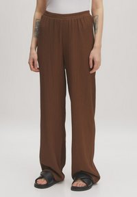 Bruine, geplooide, wijde broek met een elastische tailleband, gemaakt van een textuurstof. Draagt met zwarte slide-sandalen.