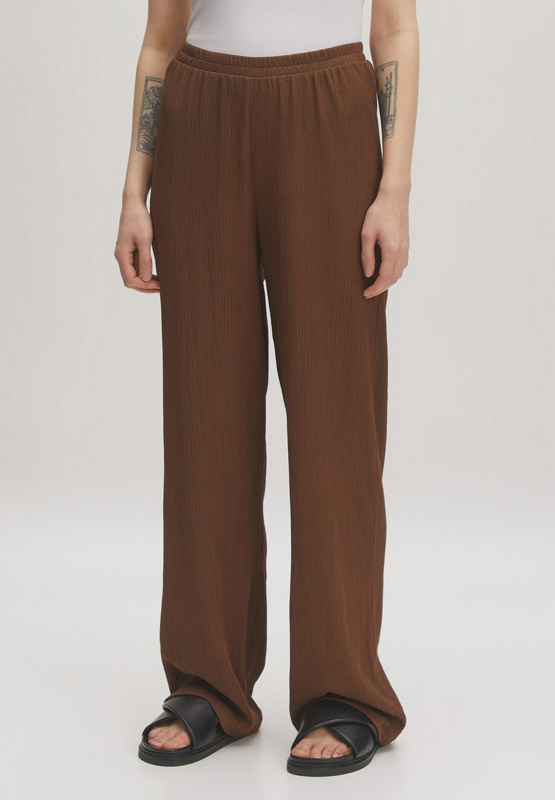 Bruine, geplooide, wijde broek met een elastische tailleband, gemaakt van een textuurstof. Draagt met zwarte slide-sandalen.