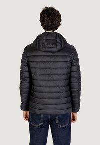 Chaqueta de abrigo negra con capucha y acolchado visible. La tela tiene una textura suave. Llevada con jeans azules, mostrada desde la parte de atrás.