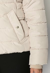 Beige quiltad jacka med ärm och ficka med tryckknapp över svarta byxor, som visar en hand och en beige mudd under ärmen.