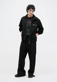 Bărbat cu păr creț, purtând ochelari transparenți, jachetă neagră cu fermoar, tricou cu imprimeu, pantaloni negri largi și pantofi negri, stând cu mâinile în buzunare.