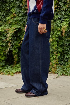 Dunkelblaue, weit geschnittene Jeans aus Denim mit sichtbaren Steppnähten, kombiniert mit einem strukturierten Pullover und braunen Slippern, stehend auf einer gepflasterten Fläche.
