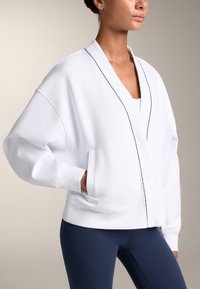 Bela oversized majica s kapuco in V-izrezom, stranskimi žepi in kontrastnim šivom na sprednji strani. Mehka tkanina, sproščen kroj in dolgi rokavi.