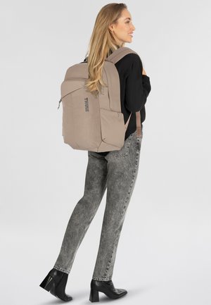 Mochila Thule gris con un acabado textil suave, compartimento principal con cremallera y bolsillos laterales. Cuenta con correas ajustables y un diseño elegante.