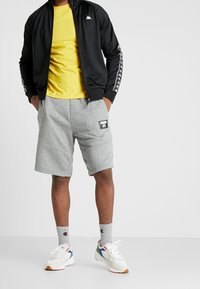 Mann trägt ein gelbes T-Shirt, eine schwarze Zip-Jacke mit Logos auf den Ärmeln, graue Shorts mit einem Aufnäher, graue Champion-Socken und weiße Turnschuhe.