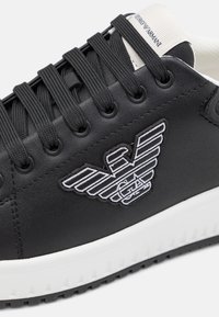 Czarne skórzane sneakersy z czarnymi sznurowadłami, białą podeszwą i białym haftowanym logo orła po boku.