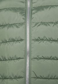 Grüner gepolsterter Weste mit einem zentralen silbernen Reißverschluss, strukturiertem Stoff mit horizontalen Nahtlinien und glatter, glänzender Oberfläche.