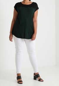 Parte superior negra y verde con un escote de encaje scalloped, combinada con jeans ajustados blancos y sandalias negras de tacón bloque. Textura de tela suave.