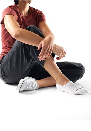 Person siddende med korslagte ben iført en rød skjorte, sorte bukser, en hvid sneaker og én hvid ankelsok på en hvid baggrund.