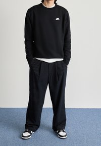 Svart hoodie med vit Nike-logotyp, kombinerad med mörka stripade byxor och svart-vita sneakers. Avslappnad passform, enkel design.