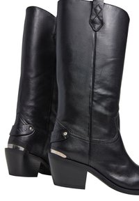 Bottes noires en cuir haute jusqu'au genou avec un talon empilé, dotées d'une sangle décorative à la cheville et d'un accent métallique sur le talon. Texture lisse, design structuré.