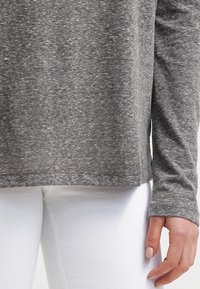 Maglietta grigia a maniche lunghe con vestibilità rilassata, texture melange e orlo arrotondato, abbinata a pantaloni bianchi. Nessun motivo o dettaglio visibile.