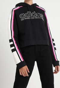 Černá zkrácená mikina s kapucí, s růžovými a bílými pruhy na rukávech, s logem a textem "adidas". Vyrobena z měkkého materiálu, volný střih.