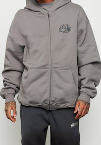 Sudadera con cremallera en color gris claro, con un logo en la parte delantera, dos bolsillos frontales, puños y cintura elásticos. Tejido suave, corte relajado y capucha.