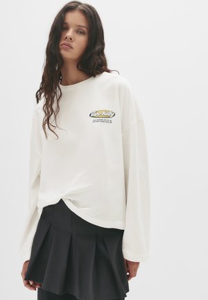 Tricou alb cu mâneci lungi, cu o croială lejeră și un grafic în față care prezintă textul "trăiește complet". Asortat cu o fustă mini neagră plisată.