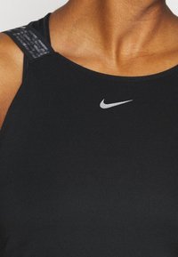Czarny sportowy top Nike z srebrnym logo, gładką fakturą i dopasowanym krojem z kontrastowymi detalami na paskach na ramionach.