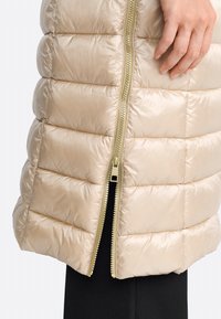 Beige Steppweste mit Reißverschluss, glänzender Oberfläche und horizontalem Steppmuster. Goldfarbener Reißverschlussdetail an der Vorderseite.