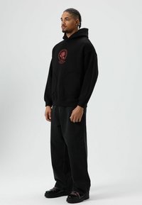 Zwarte hoodie met zak aan de voorkant en rode grafische details, gecombineerd met losse zwarte jeans en zwarte schoenen met roze veters.