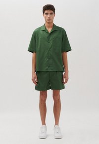 Camicetta verde a maniche corte con tasca e pantaloni corti abbinati, realizzati in un tessuto morbido, abbinati a sneaker bianche su uno sfondo neutro.