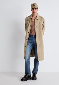 Abrigo beige con botones, camiseta de tirantes marrón, jeans azules deslavados y zapatos de plataforma negros. Combinado con una gorra beige a juego.