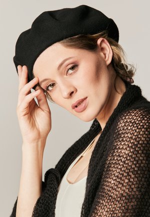 Czarny wełniany beret noszony lekko na skos, w połączeniu z czarnym, luźno dzierganym szalem. Modelka ma na sobie neutralną bluzkę i delikatny makijaż.