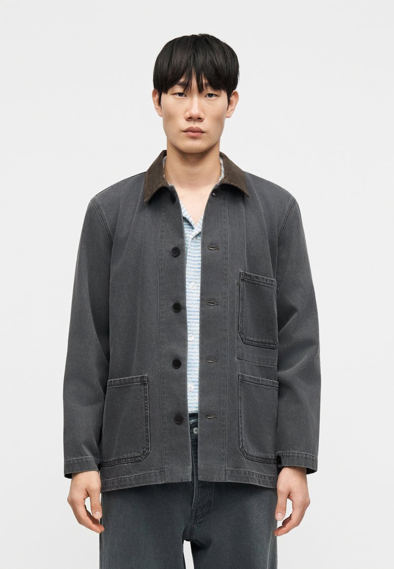 Γκρι denim μπουφάν με καφέ γιακά από κορδόνι, κλείσ ιμο με κουμπιά μπροστά, δύο μεγάλες κάτω τσέπες και άνετη γραμμή. Φοριέται πάνω από ριγέ πουκάμισο.