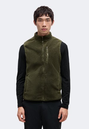 Un homme se tient face à l’objectif devant un fond uni. Il porte un gilet polaire olive sur un haut noir à manches longues et un pantalon noir.