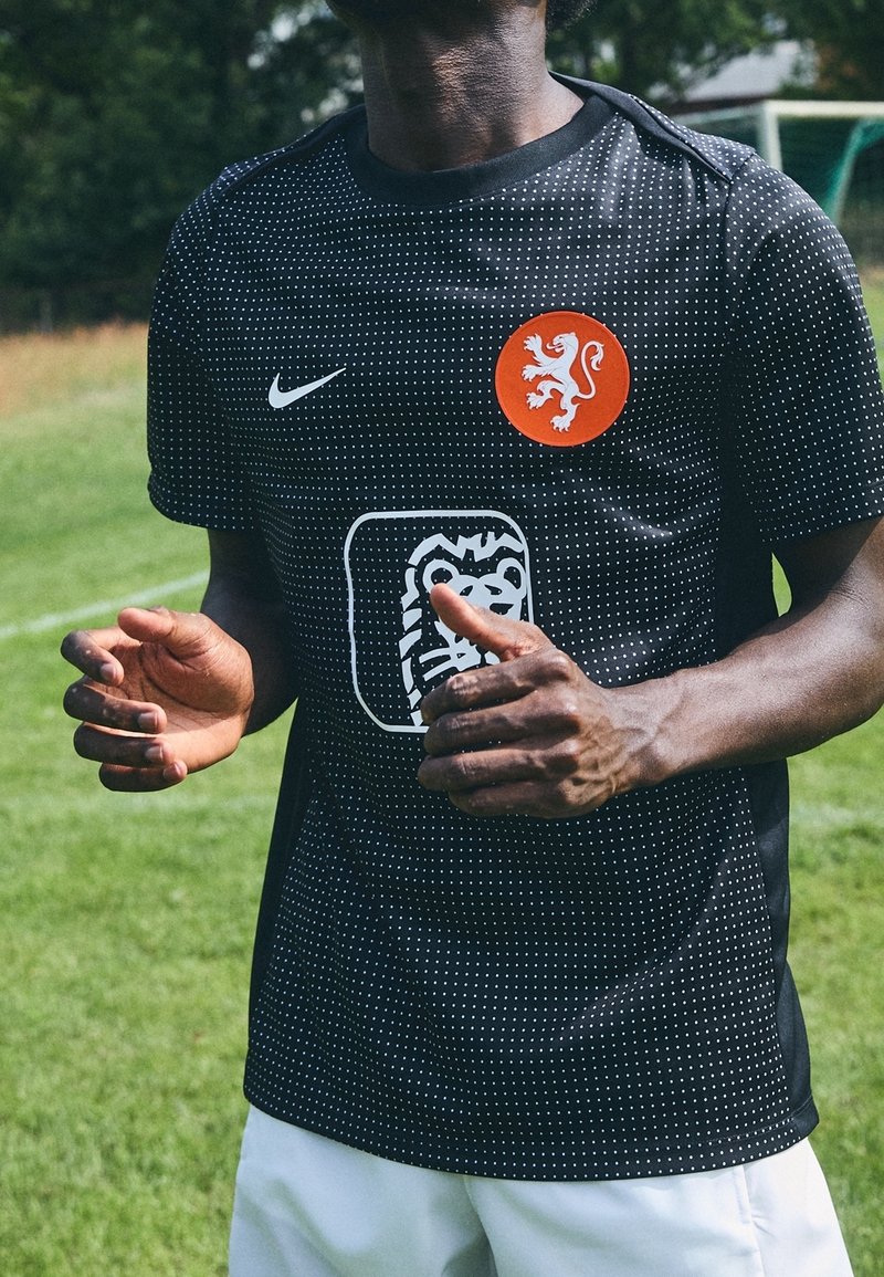 Svart tröja med vita prickmönster, Nike-logga och en orange lejonemblem på bröstet, tillverkad av ventilerande tyg.